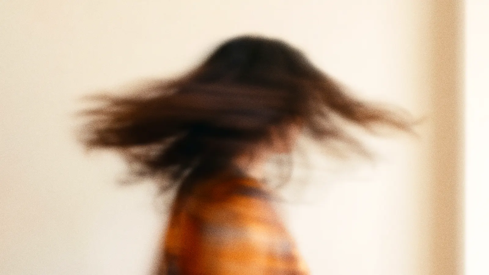 Una mujer gira la cabeza deprisa ante una pared clara; su pelo y su camisa naranja se convierten en estelas de movimiento